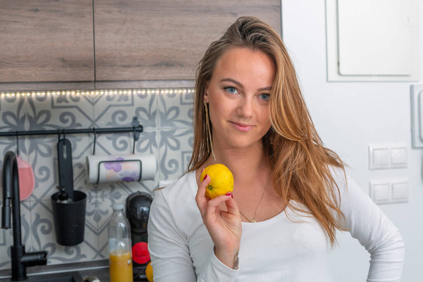 beautiful young blue-eyed blonde in the kitchen holding a lemon in her hand looking directly at the camera - Fotografie, Obrázek