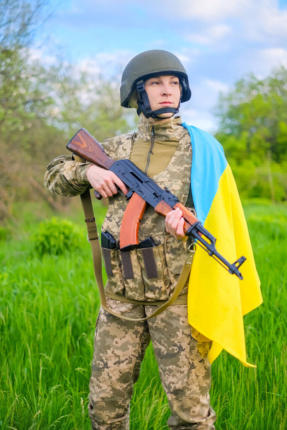 Kamuflaj üniforması ve miğferi olan, Ukrayna bayrağı omuzlarıyla çevrili ve başka yöne bakan silahlı, kararlı kadın asker. Ukraynalı kadın vatansever. Dikey yönelim - Fotoğraf, Görsel