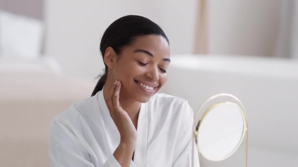 Juventud y belleza. De cerca retrato de la joven mujer afroamericana bonita mirando el espejo en el baño y preening, sonriendo a sí misma, acariciando su cara - Imágenes, Vídeo