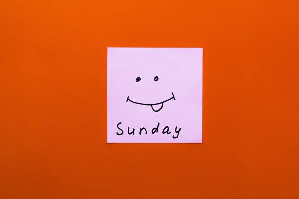 vrchní pohled na papír s nedělním nápisem a veselým úsměvem s vystrčeným jazykem na oranžovém pozadí - Fotografie, Obrázek