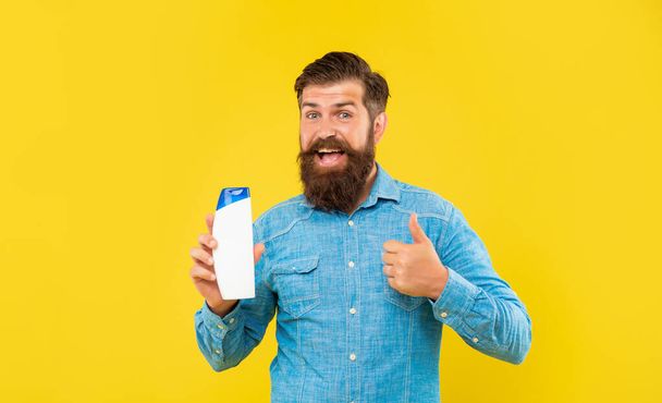 amazed brutal bearded man hold shampoo bottle on yellow background, skincare. thumb up. - Foto, afbeelding