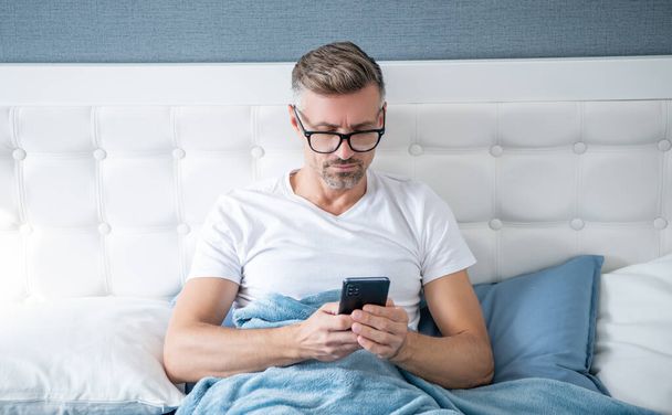 mature man in bed check email on phone wearing eyeglasses. - 写真・画像