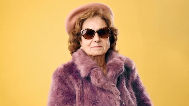 Elegante mujer mayor en gafas de sol con un abrigo de piel y una boina mientras posa sobre un fondo amarillo. - Metraje, vídeo