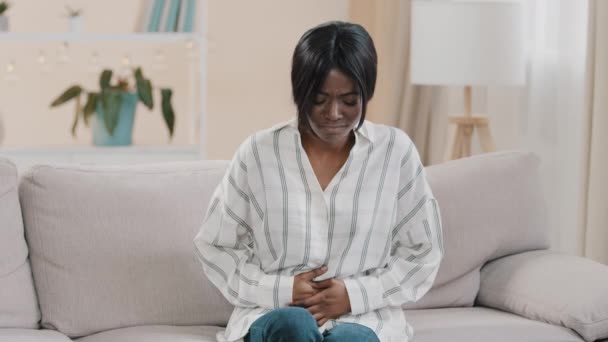 Young upset unhealthy sad african american woman sitting in room on couch holding hands on stomach suffering from abdominal pain feeling spasm from diarrhea menstrual painful feelings symptom nausea - Footage, Video