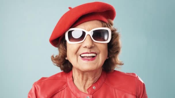 Elegante mujer mayor con gafas de sol mirando a la cámara y empezar a reír mientras está de pie sobre un fondo aislado. - Metraje, vídeo