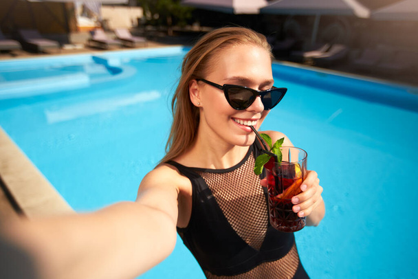 Travel blogger woman in bikini taking selfie photo with cocktail near swimming pool. Pov - lifestyle vlogger selfie from vacation on luxury resort. Female in swimwear drinks beverage on tropical spa. - Foto, imagen