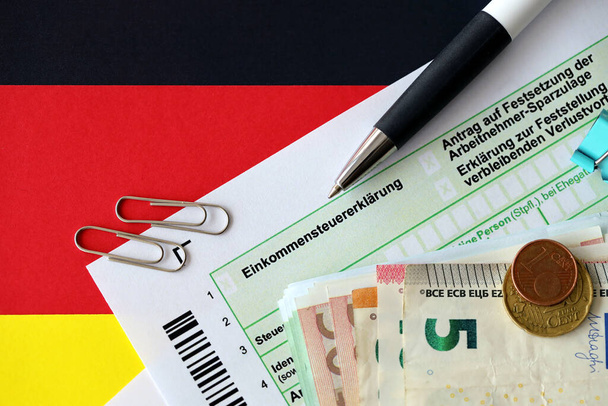 Německý formulář přiznání k dani z příjmu s perem a eurobankovkami na peníze leží na vlajce zblízka. Daňoví poplatníci v Německu, kteří k placení daní používají jako hlavní měnu euro - Fotografie, Obrázek