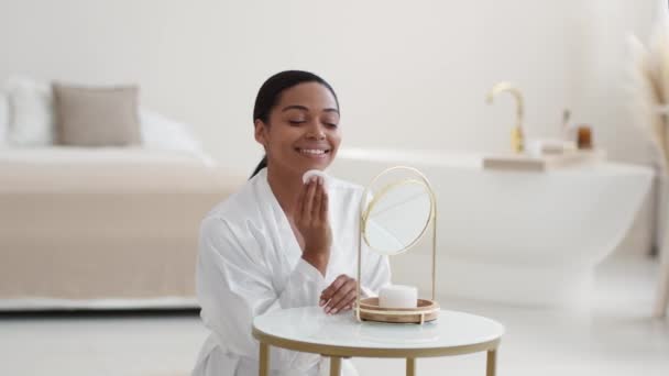 Concepto de limpieza de la piel. Joven dama afroamericana feliz limpiando su cara con almohadilla de algodón y loción, mirando al espejo en el baño, cámara lenta, espacio libre - Imágenes, Vídeo