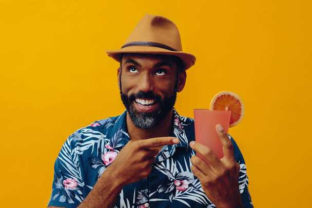 handsome african american man wearing hawaian shirt and hat with orange juice posing on yellow background - Fotografie, Obrázek