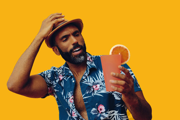 handsome african american man wearing hawaian shirt and hat with orange juice posing on yellow background - Foto, immagini