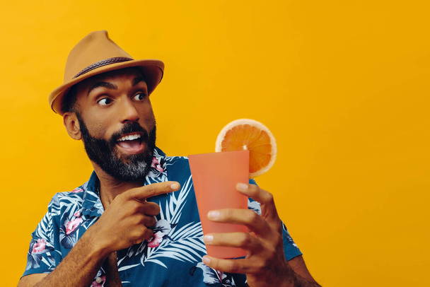 handsome african american man wearing hawaian shirt and hat with orange juice posing on yellow background - Photo, Image