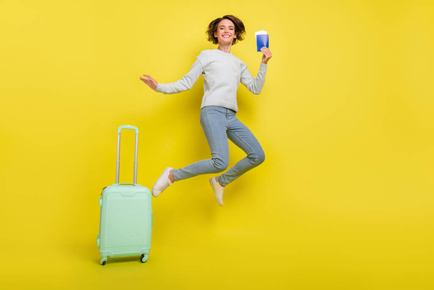Full body photo of funky millennial relax lady jump with bag passport wear pullover jeans sneakers isolated on yellow background. - Fotografie, Obrázek