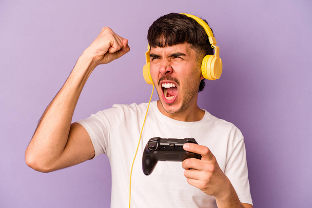 Young hispanic man playing with a video game controller isolated on purple background raising fist after a victory, winner concept. - Photo, image