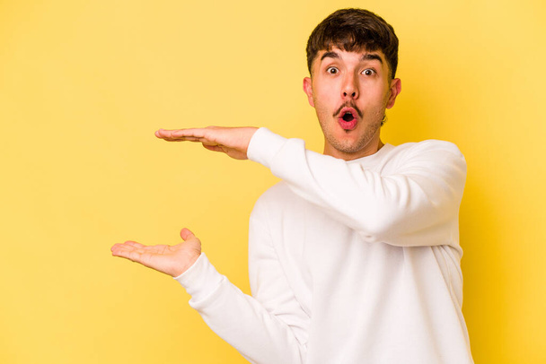 Young caucasian man isolated on yellow background shocked and amazed holding a copy space between hands. - Valokuva, kuva