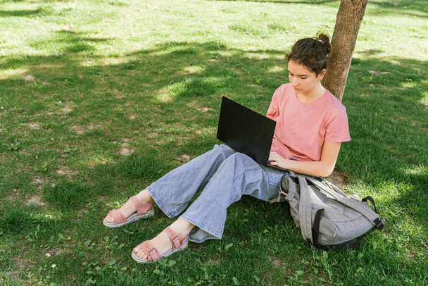 teenage girl, student or student of gymnasium, sits on green lawn in park with laptop, does homework, writes essay, completes online assignments. Use of modern technologies and gadgets by teenagers - Photo, image