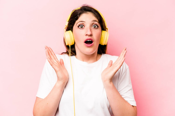 Joven mujer caucásica escuchando música aislada sobre fondo rosa sorprendida y conmocionada. - Foto, Imagen