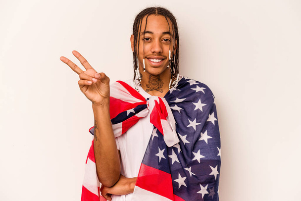 Junger afroamerikanischer Mann mit einer amerikanischen Flagge auf weißem Hintergrund, die fröhlich und unbeschwert ein Friedenssymbol mit Fingern zeigt. - Foto, Bild