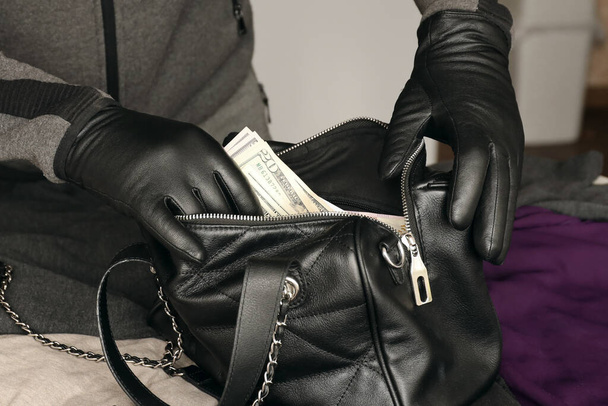 Robber in black outfit and gloves see in opened stolen women bag. The thief takes out US dollar bills from a womans handbag in kitchen interior - Foto, imagen