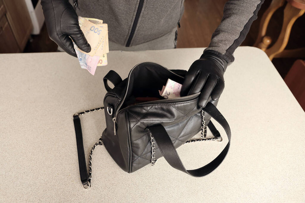 Fekete ruhás rabló és kesztyű a nyitott lopott női táskában. A tolvaj előveszi az ukrán pénzt egy női kézitáskából a konyha belsejében. - Fotó, kép