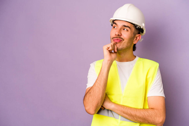 Jeune travailleur hispanique homme isolé sur fond violet regardant latéralement avec une expression douteuse et sceptique. - Photo, image