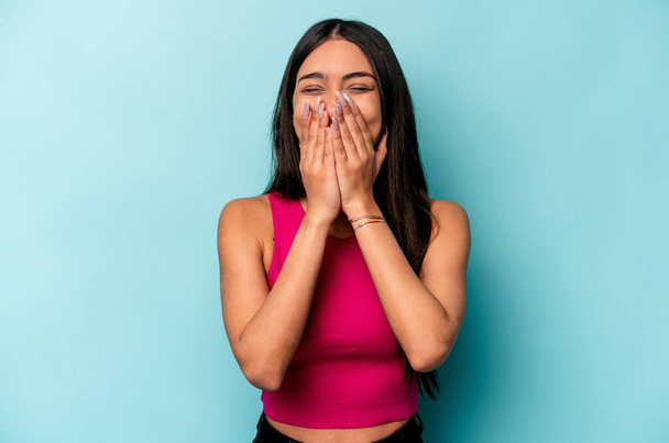 jonge Spaanse vrouw geïsoleerd op blauwe achtergrond lachen over iets, bedekken mond met handen. - Foto, afbeelding
