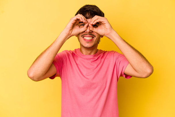 Young caucasian man isolated on yellow background showing okay sign over eyes - Foto, imagen
