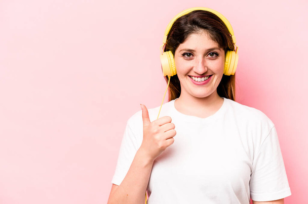 Молода біла жінка слухає музику ізольовано на рожевому фоні посміхається і піднімає великий палець вгору
 - Фото, зображення