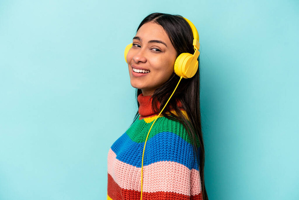 Mladá hispánka poslouchající hudbu izolovanou na modrém pozadí se dívá stranou s úsměvem, veselým a příjemným. - Fotografie, Obrázek