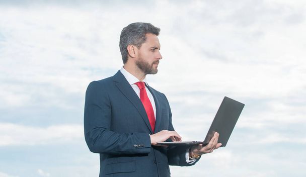 Gökyüzü arkaplanındaki dizüstü bilgisayarı kullanarak takım elbiseli CEO. - Fotoğraf, Görsel