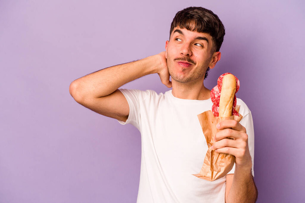 Mladá hispánka drží sendvič izolovaný na purpurovém pozadí, dotýká se týla hlavy, přemýšlí a dělá rozhodnutí. - Fotografie, Obrázek