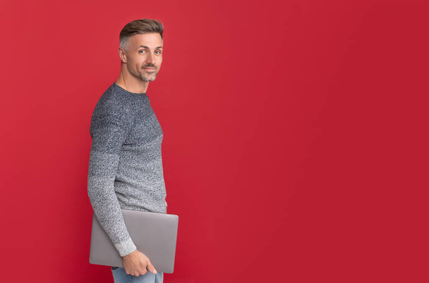 positive freelancer. grizzled guy in sweater on red background. hoary man holding laptop. computer communication. modern technology. - Photo, Image