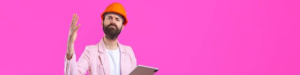 Portret młodego inżyniera budowlanego nosi pomarańczowy, twardy kapelusz, w różowej kurtce stojącej na czerwonym tle pracowni. Mężczyzna z tabliczką w rękach.. - Zdjęcie, obraz