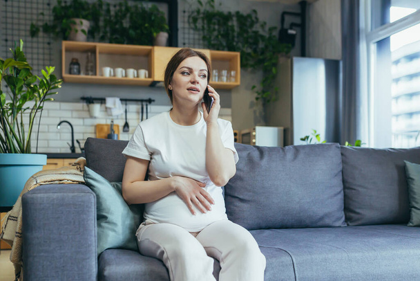 Pregnant beautiful woman sitting on the couch at home, talking on a cell phone with a doctor, calling an ambulance, giving birth - Fotografie, Obrázek