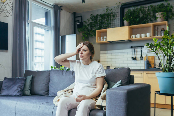 Feeling unwell. A pregnant woman is ill, has a headache, holds her hand behind her head. Sitting at home in home clothes on the couch - Фото, изображение