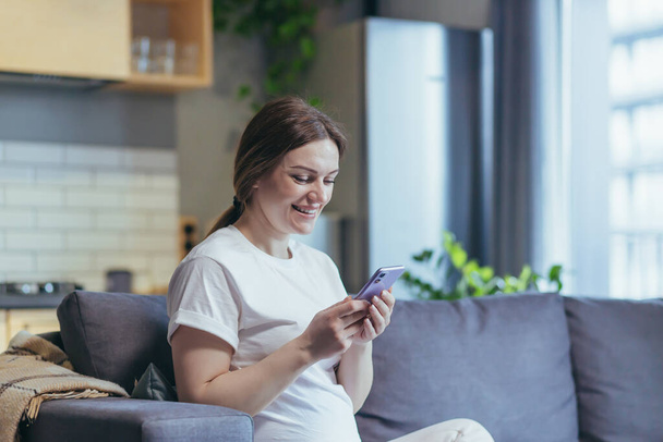 Maternity leave. Pregnant beautiful woman sitting on the couch at home, holding a mobile phone, typing messages, calling, watching, receiving good news. Smiling, happy - Fotografie, Obrázek