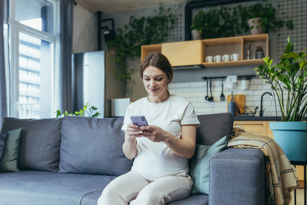 Maternity leave. Pregnant beautiful woman sitting on the couch at home, holding a mobile phone, corresponding in parent chats, typing messages, calling, watching, receiving good news. Smiling, happy - Foto, Imagem