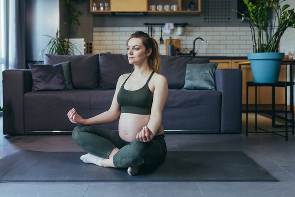 Pregnant beautiful woman doing yoga at home, sitting on a sports mat in the lotus position, closing her eyes, meditating. - Valokuva, kuva