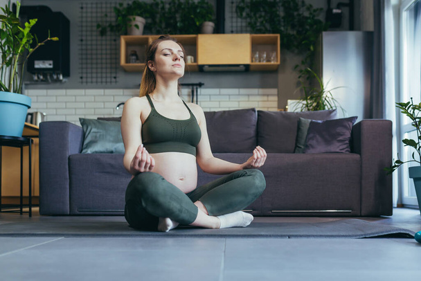 Mujer hermosa embarazada haciendo yoga en casa, sentada en una alfombra deportiva en posición de loto, cerrando los ojos, meditando. - Foto, imagen
