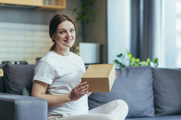 Pregnant beautiful woman received a parcel by courier home, package, gift. He sits on the sofa at home, opens a paper box, rejoices, smiles - Photo, image