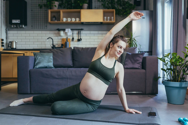 Pregnant beautiful woman goes in for sports at home, sits and lies on a sports mat in a sports uniform, does exercises, is satisfied - Fotografie, Obrázek