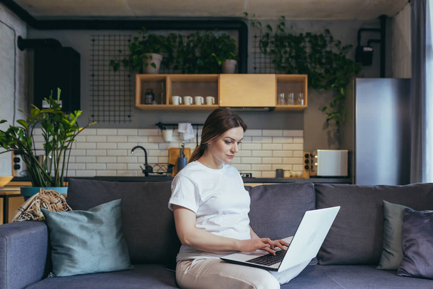 Maternity leave. Pregnant beautiful woman sitting on sofa at home with laptop in home clothes, ordering baby goods online, choosing, buying clothes and toys for baby. - Valokuva, kuva