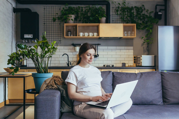 Maternity leave. Pregnant beautiful woman sitting on sofa at home with laptop in home clothes, ordering baby goods online, choosing, buying clothes and toys for baby. - Фото, зображення