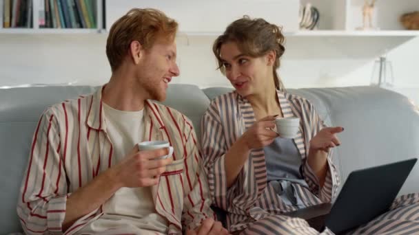 Young couple enjoy coffee on comfy couch closeup. Morning leisure time at home. Smiling beautiful family talking drinking beverages together on weekend. Relaxed girl ginger man laptop browsing web - Footage, Video