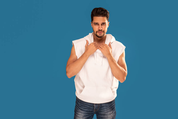 Fashionable young handsome man posing in white tank top with hood on blue studio background. Bearded guy looking at the camera. Copy space. - Φωτογραφία, εικόνα
