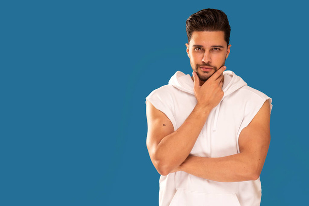 Fashionable young handsome man posing in white tank top with hood on blue studio background. Bearded guy looking at the camera. Copy space. - Valokuva, kuva