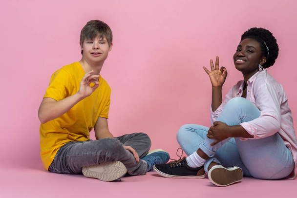 Good mood. Dark-skinned woman and caucasian guy with down syndrome smiling showing hand signs at camera sitting on floor - Фото, изображение