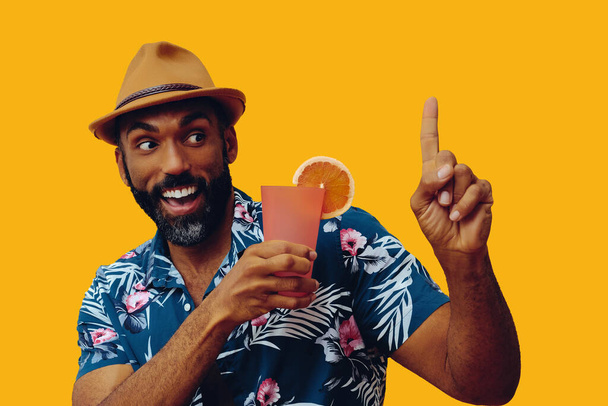 Havai gömleği ve sarı arka planda portakal suyu pozu veren şapkalı yakışıklı Afro-Amerikan bir adam. - Fotoğraf, Görsel