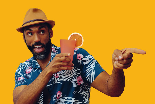 handsome african american man wearing hawaian shirt and hat with orange juice posing on yellow background - Foto, imagen