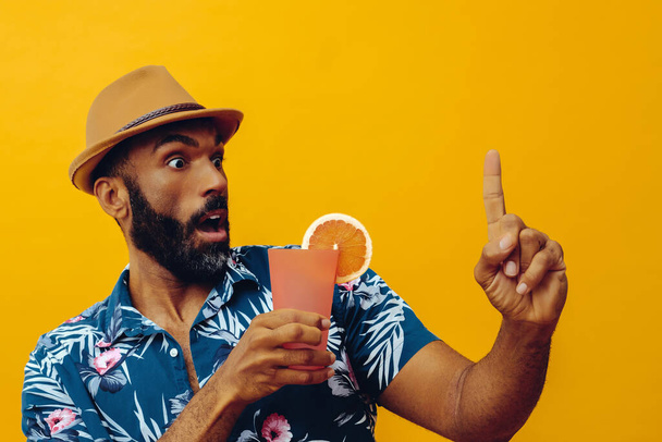 handsome african american man wearing hawaian shirt and hat with orange juice posing on yellow background - Fotoğraf, Görsel