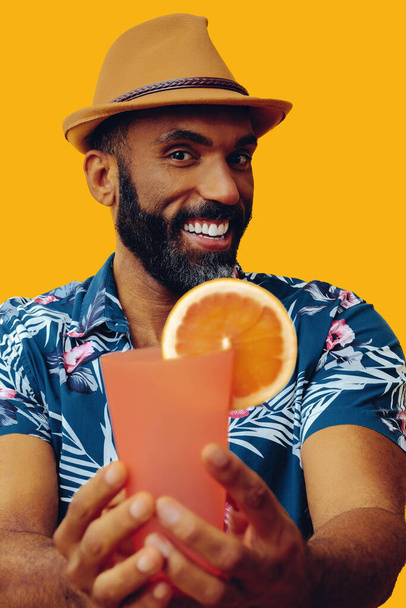 hombre afroamericano guapo vistiendo camisa hawaiana y sombrero con jugo de naranja posando sobre fondo amarillo - Foto, Imagen